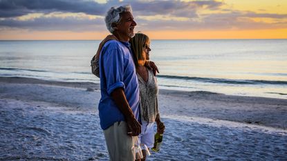 An older couple stand arm in arm on the beach and watch the sun set.