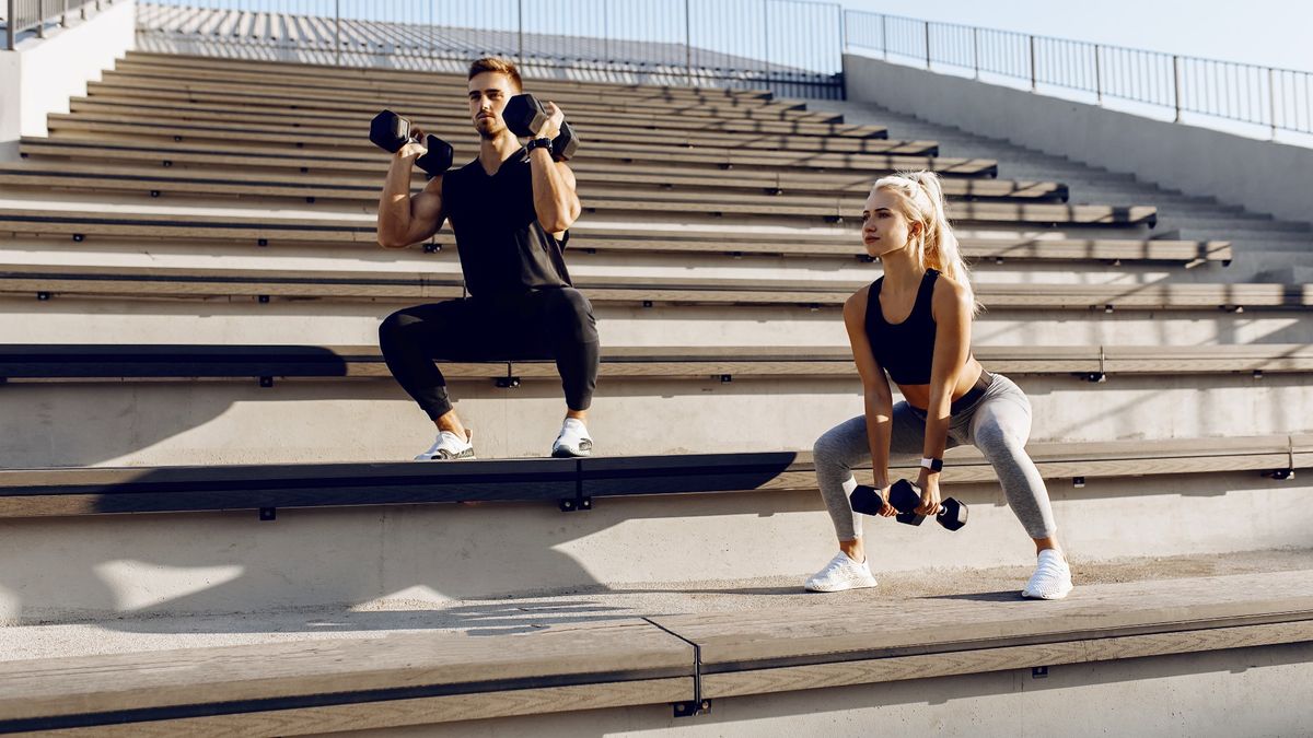 Man and woman outdoors on steps performing dumbbell squat variations in the sunshine. Man in front rack squat and woman in sumo squat