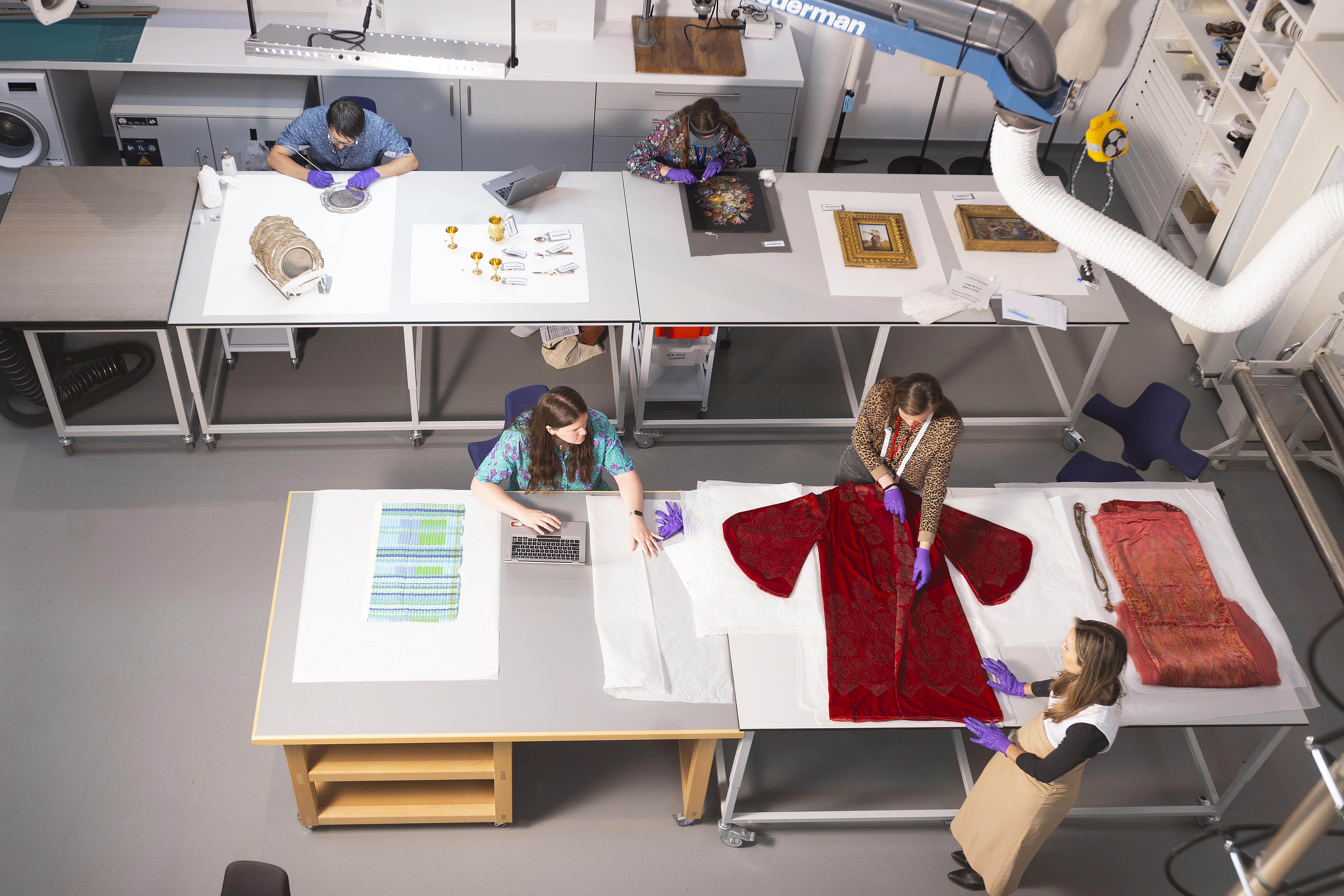 A birds-eye view of two workstations with a red dress, artwork in golden gilt frames and other artefacts being handled by visitors
