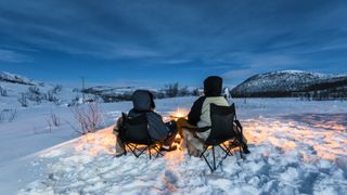 Two people sit in front of a campfire in the snow