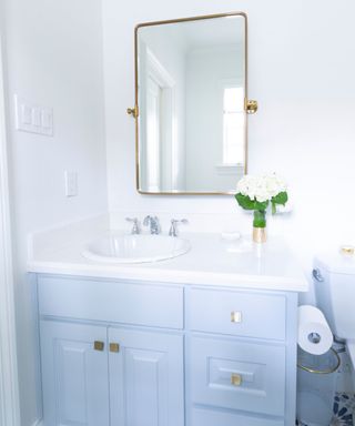 A white bathroom with a gold rectangular tilt mirror, a light blue vanity with a white sink and countertop with white flowers on it