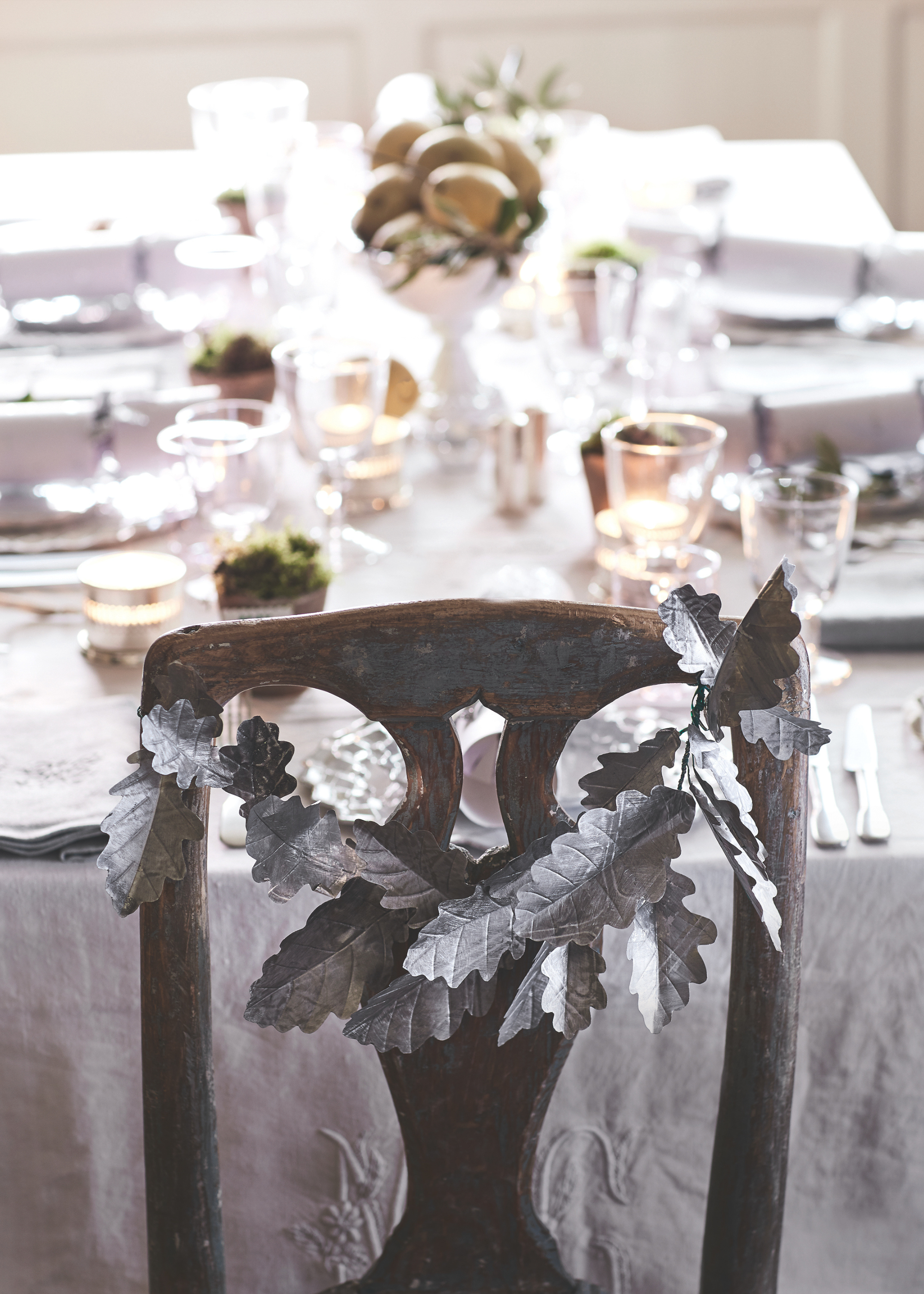 A dining chair is adorned with a silver-coated leaf garland in front of a table set for Christmas with a white tablecloth and fruit in a bowl in the center