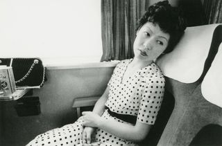 Black and white portrait of a Japanese woman in a white dress leaning back into a seat on a train