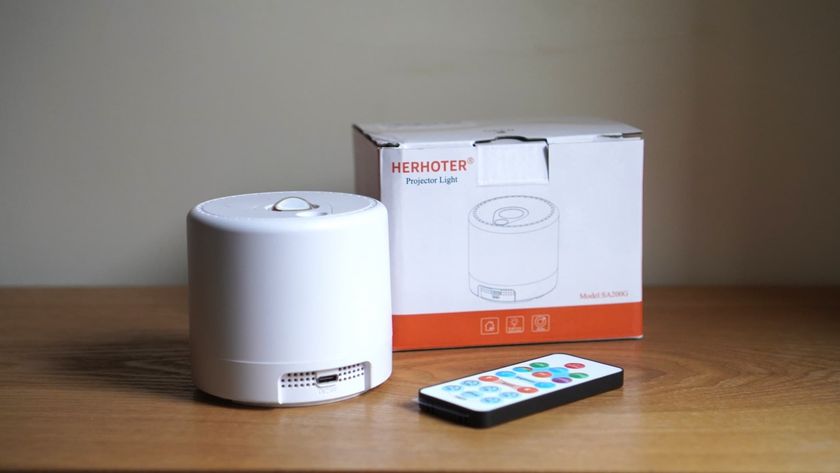 A white spherical projector is placed next to a multicolor remote control and a white box, all on a brown wooden surface.