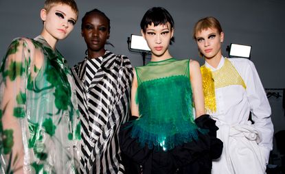Models wear green floral dress, striped black and white dress, green top with feather, and white shirt and trousers
