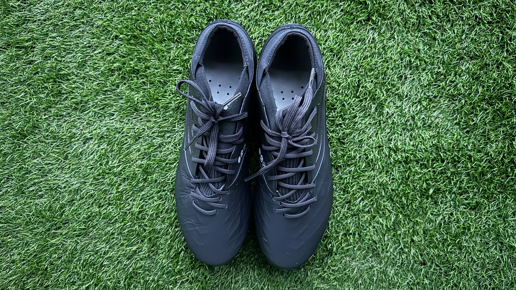Two black football boots, both with a black sole and black laces sit side by side on green artificial grass. Kipsta Viralto IV Pro