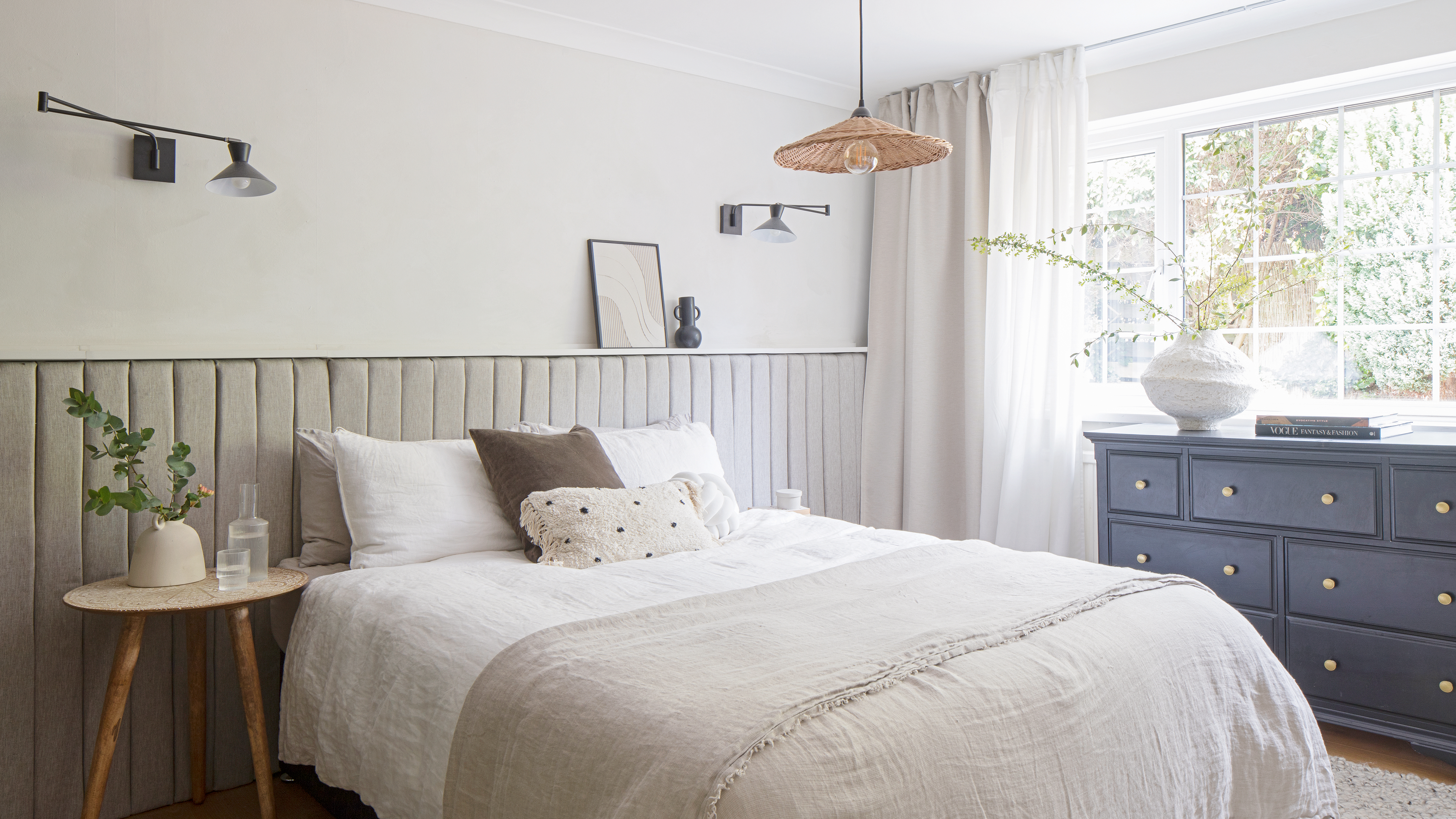 Neutral bedroom with grey panelled half height wall and bed dressed with white linen and a dark grey chest of drawers under the window