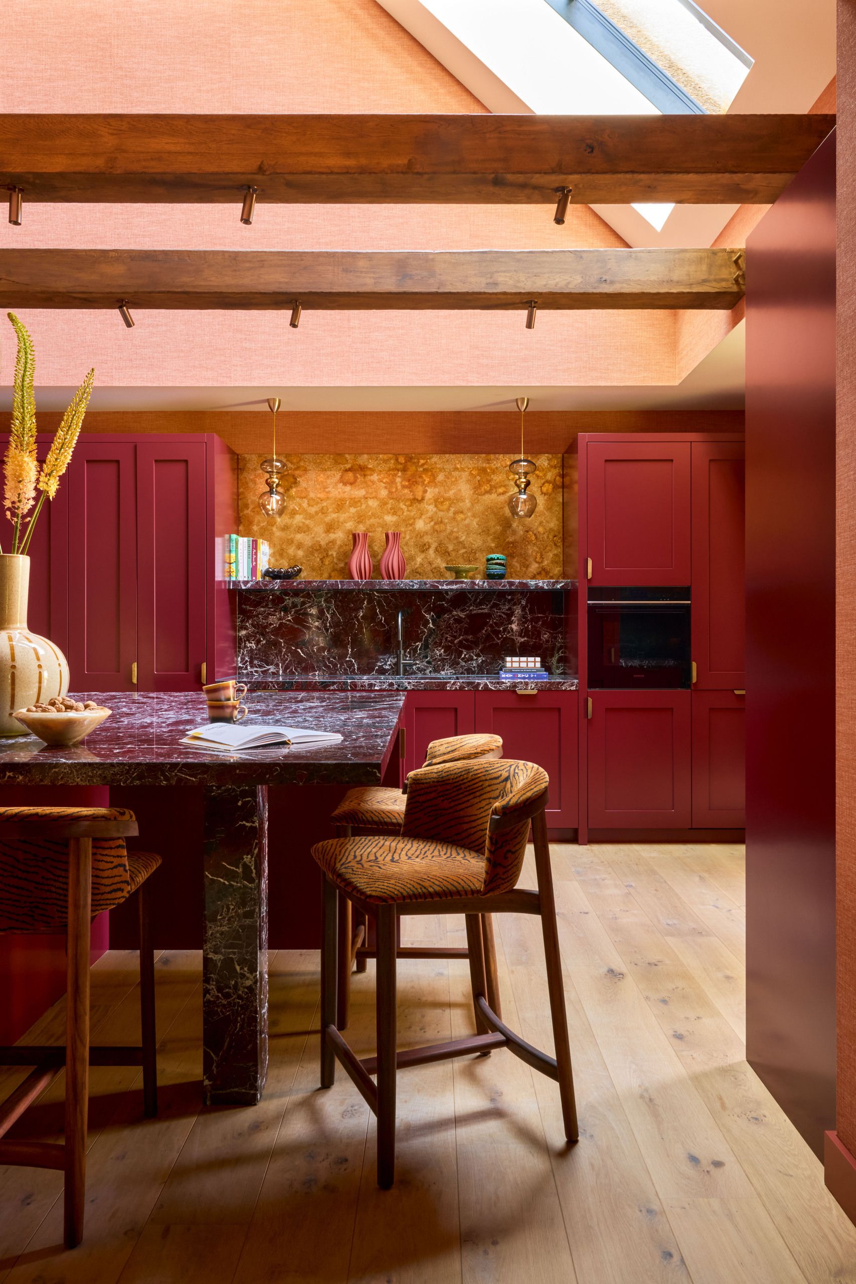 Image of a red kitchen with red cabinetry, pink vaulted ceilings, a skylight, and a red marble island with tiger-print bar chairs around it.