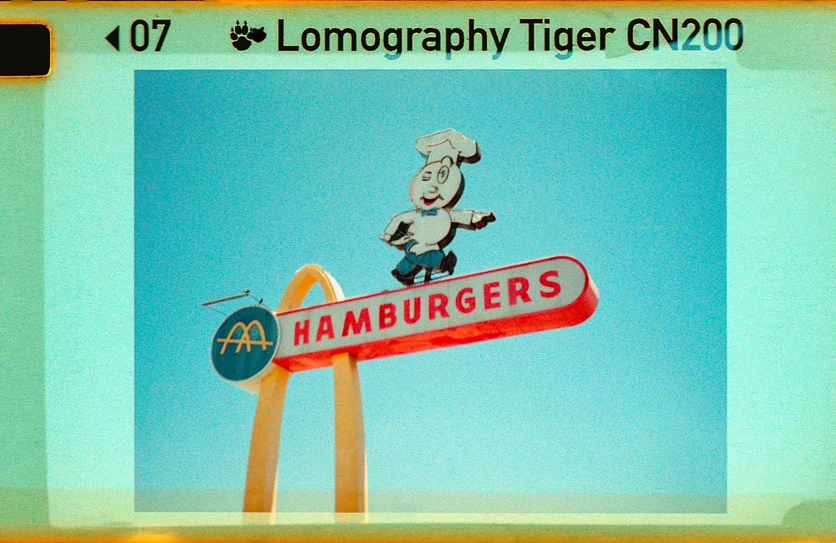 Vintage photo of a retro "HAMBURGERS" sign with a chef figure and golden arches against a clear blue sky.