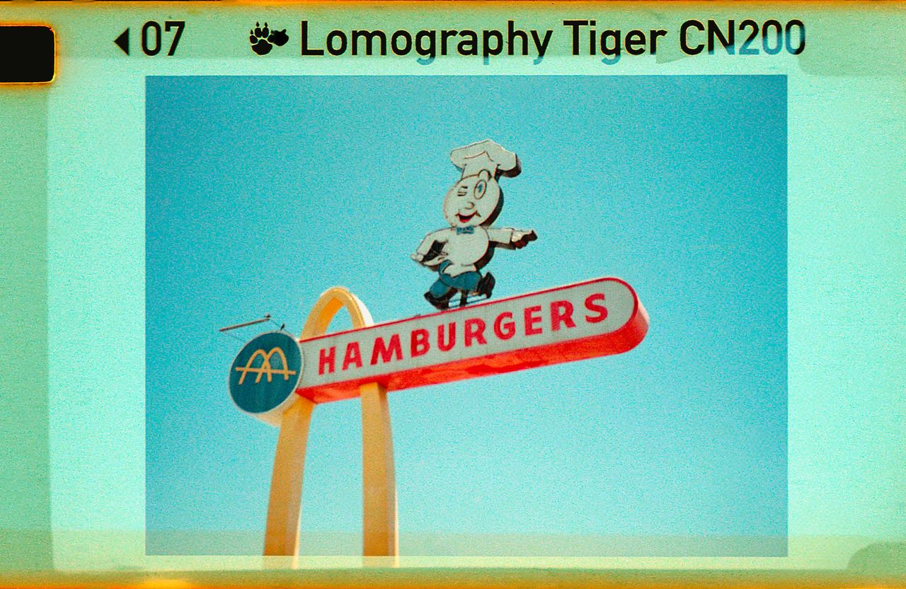 Vintage photo of a retro "HAMBURGERS" sign with a chef figure and golden arches against a clear blue sky.