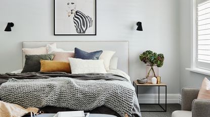 A main bedroom with black wall sconce fixtures on either side of bed, framed wall art above bed, grey wall paint decor, wood and metal side table, grey carpet on floor, and assortment of bedding including chunky grey throw, pillows and scatter cushions