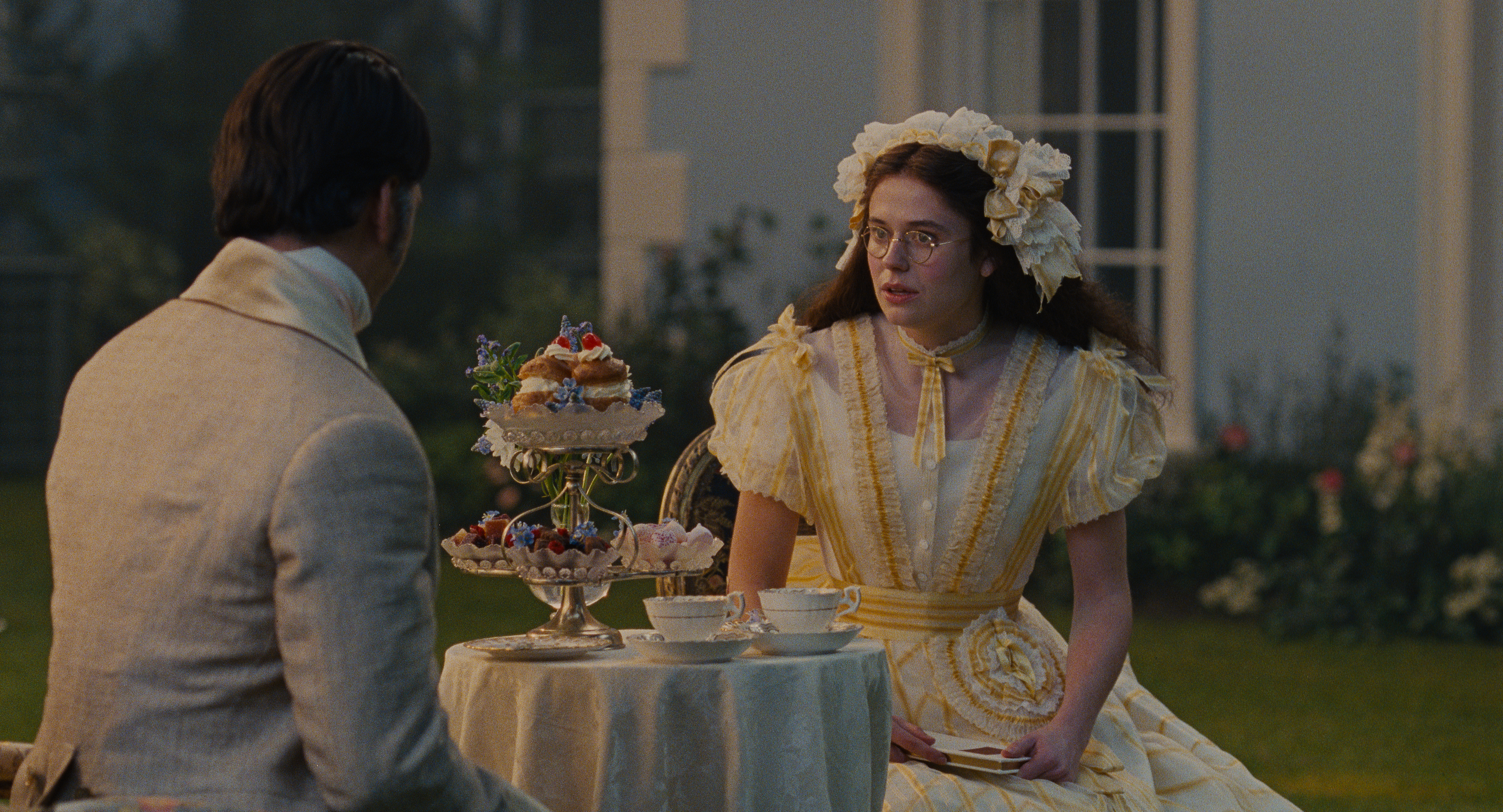 alison oliver as isabella wearing a yellow and white frilly gown with a bonnet covered in bows talking to shazad latif as edgar linton at a tea party table outside in a still from wuthering heights