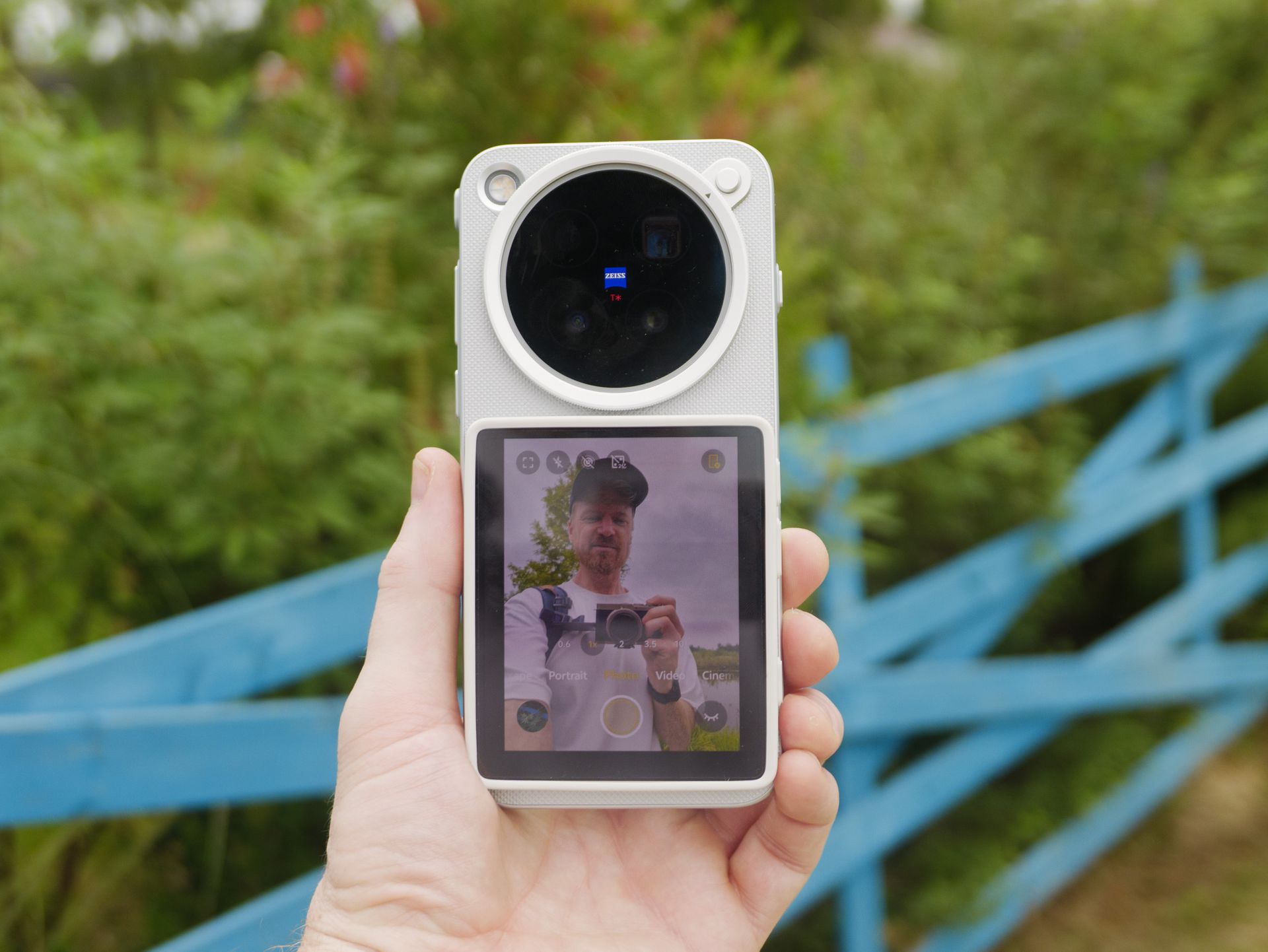 The Vivo X500 Pro Max phone in user's hand, in its dedicated case for magnetic accessories, with Vivo's selfie screen attached, and person is vlogging by a blue fence and greenery