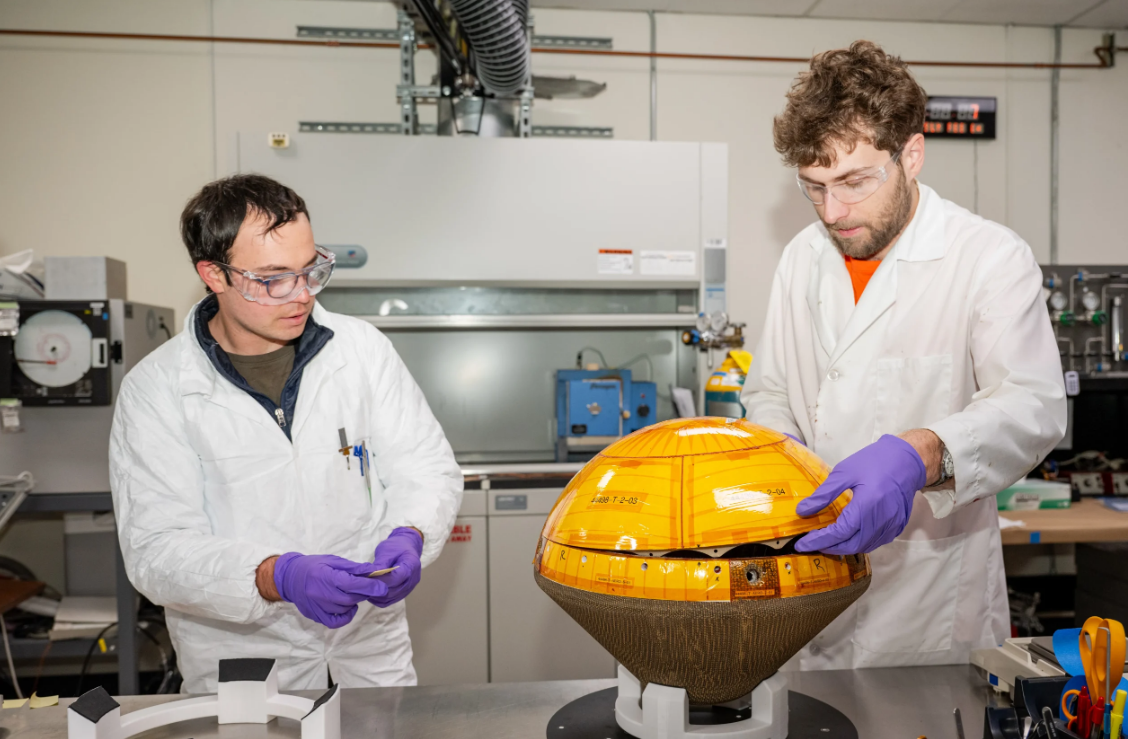 Two scientists in lab coats place an orange dome on a cone-like spacecraft the size of a large remote-controlled car