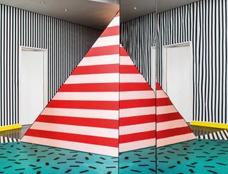 Abstract installation featuring red and white stripped pyramid, black and white stripped background and teal patterned floor