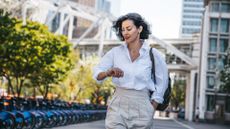 Stylish woman on 15-minute walk wearing shirt and trousers with bag, looking at watch in the city