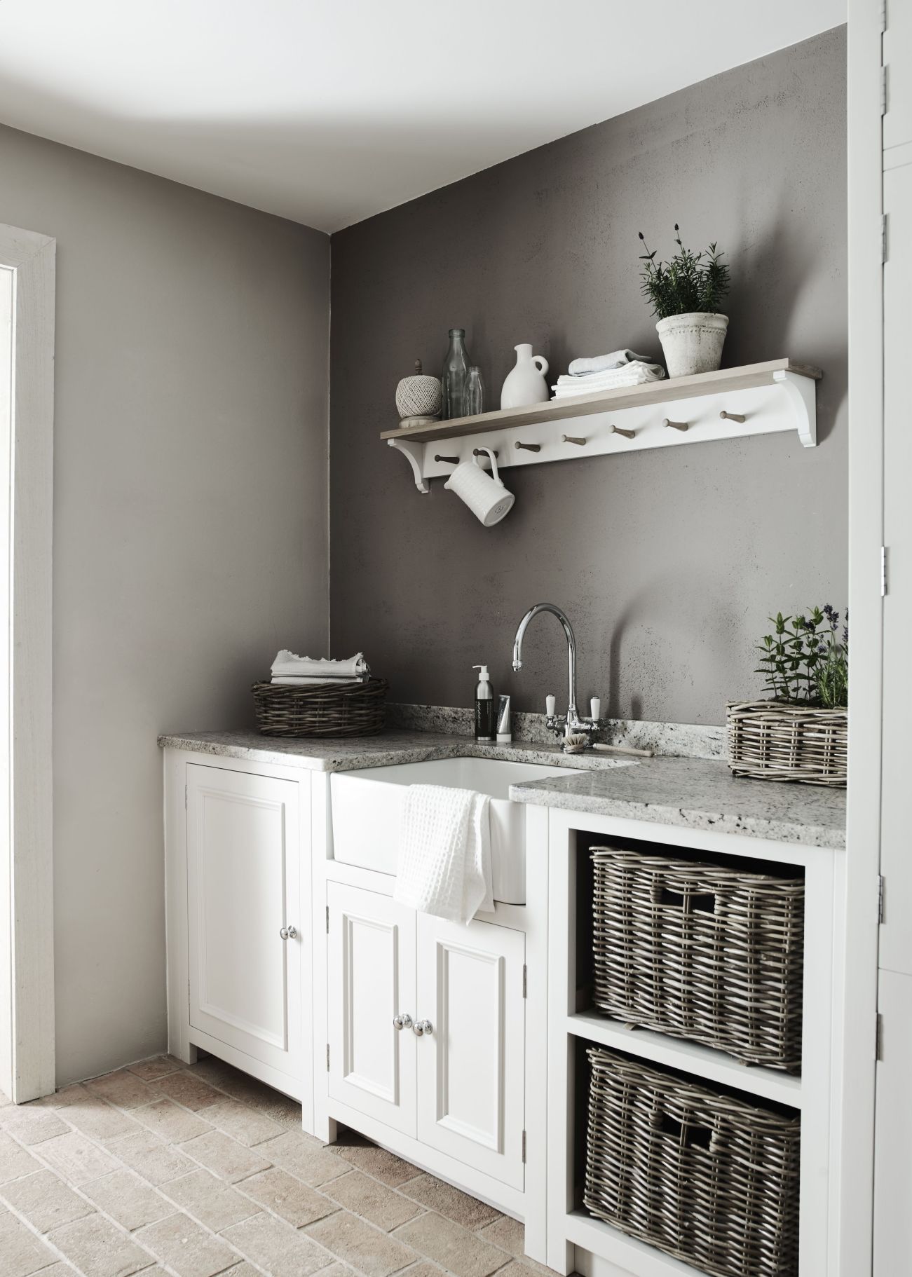 A pale gray laundry room with brick flooring, white cabinetry, wicker baskets for storage and a white wooden wall shelf.