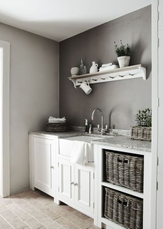 A pale gray laundry room with brick flooring, white cabinetry, wicker baskets for storage and a white wooden wall shelf.