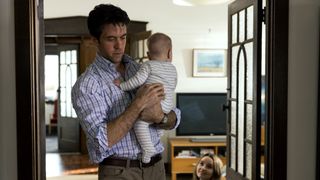 Ashley Zukerman as Belle's partner Clive, looking downcast and holding her baby son, while she looks on.