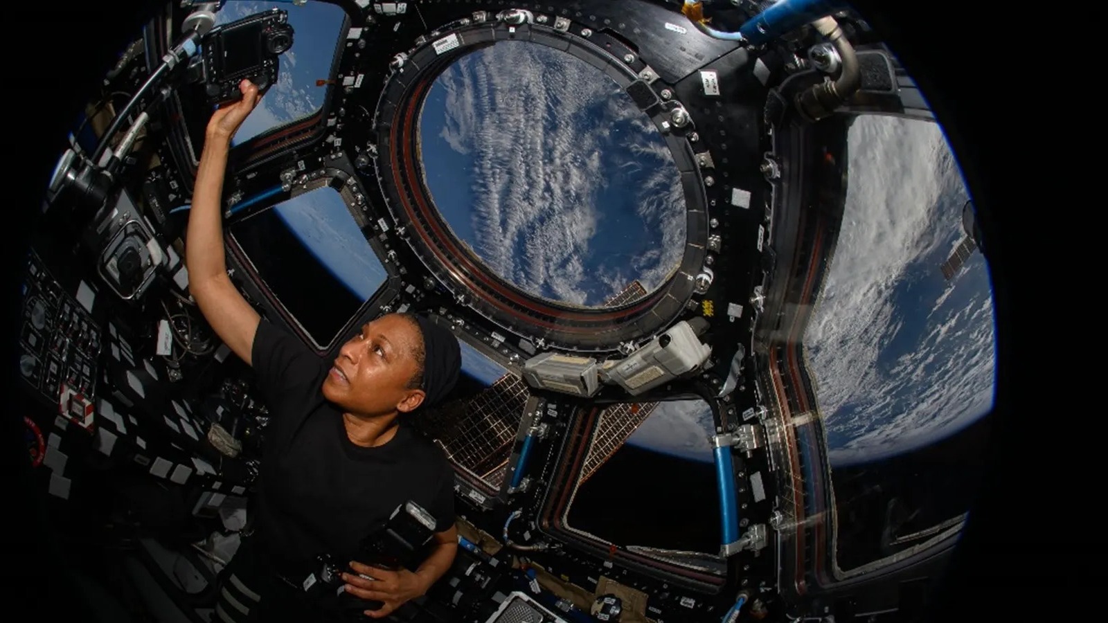 an astronaut in the cupola with many windows behind her. she raises a camera in front of the window and faces the screen of the camera