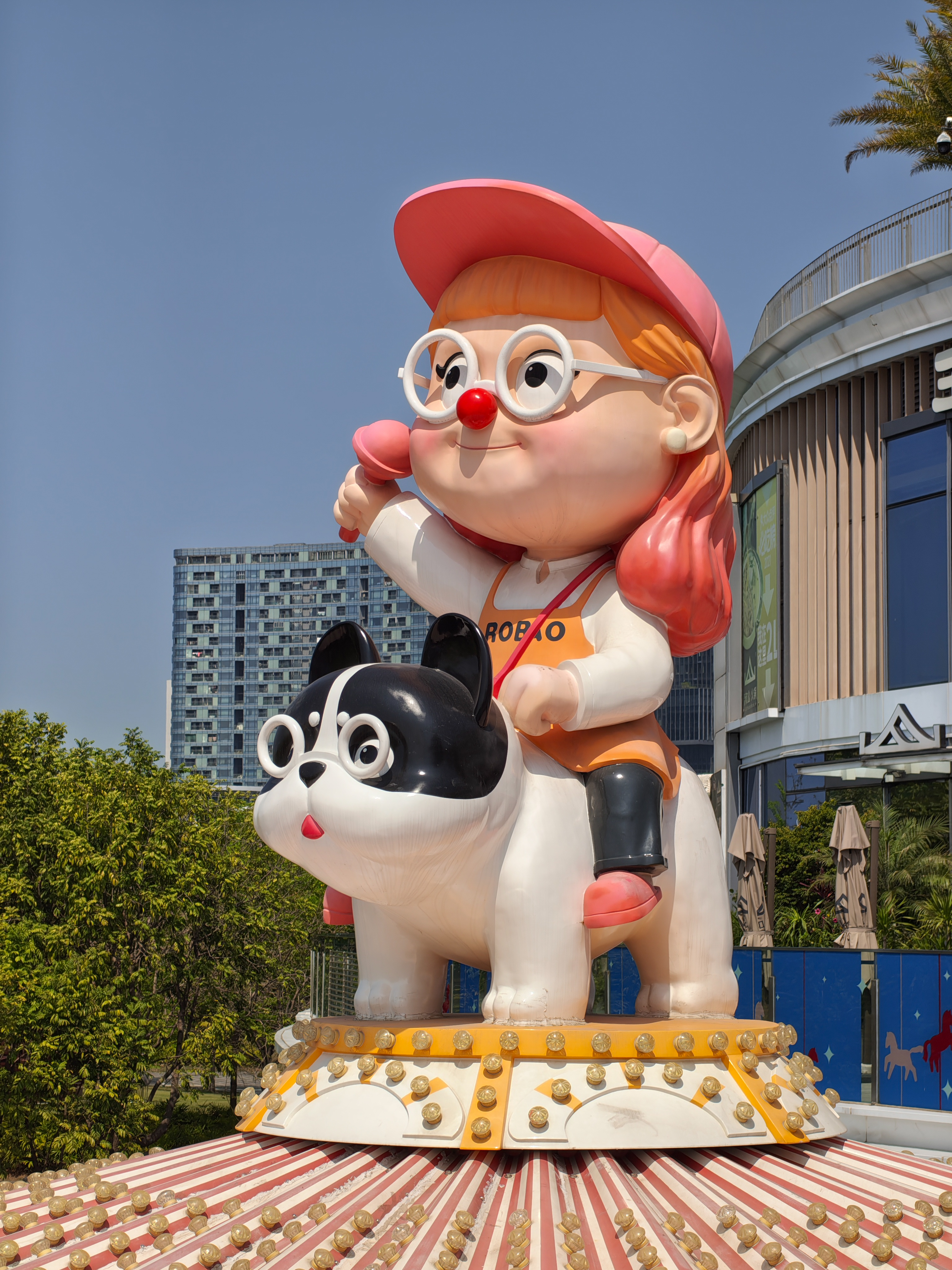 A large, colorful outdoor statue of a cartoon girl with orange hair, a pink cap, and round white glasses. She is sitting on the back of a large black-and-white French Bulldog statue. The entire display is mounted on a carousel-style base with small lightbulbs. A modern glass building and green trees are visible in the background.