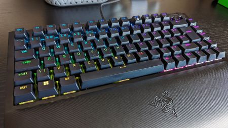 A black gaming keyboard with RGB backlit keys on a desk. Keys display a gradient of colors, creating a vibrant and dynamic effect. Razer logo visible.
