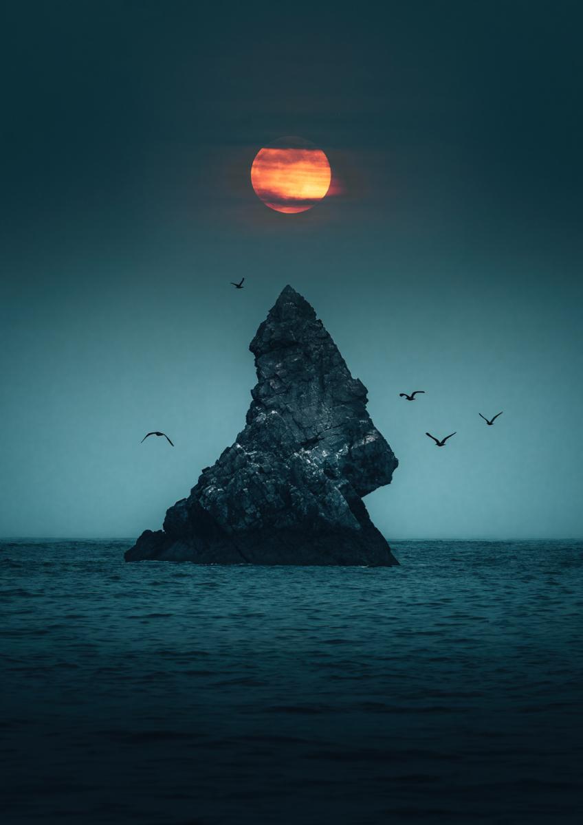A large, sharp rock just out of the sea as gulls fly around on either side. Above it, an orange full moon hangs peering between hazy blue/gray clouds.