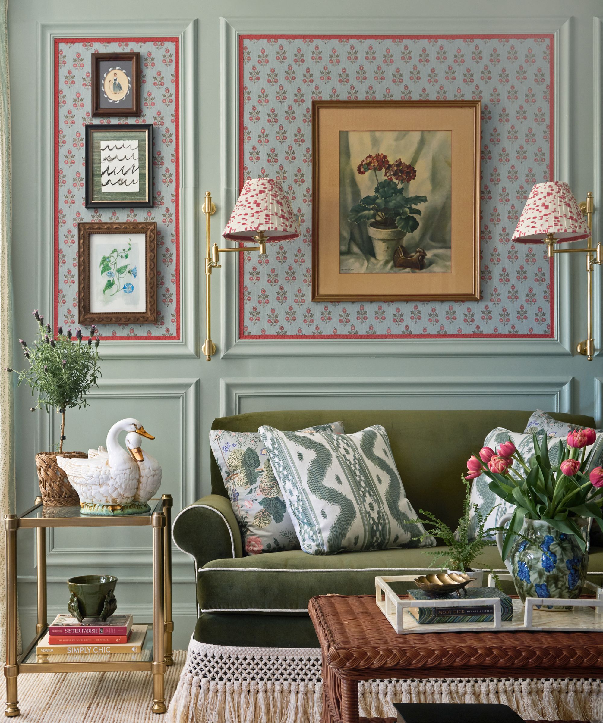 Living room with pale blue walls, vintage artwork, dark green velvet sofa with white piping and fringe, rattan coffee table, and gold side table with swan planters