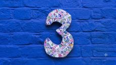 The number 3, made up of a collage of colorful broken pottery, is mounted on a brick wall painted bright blue.