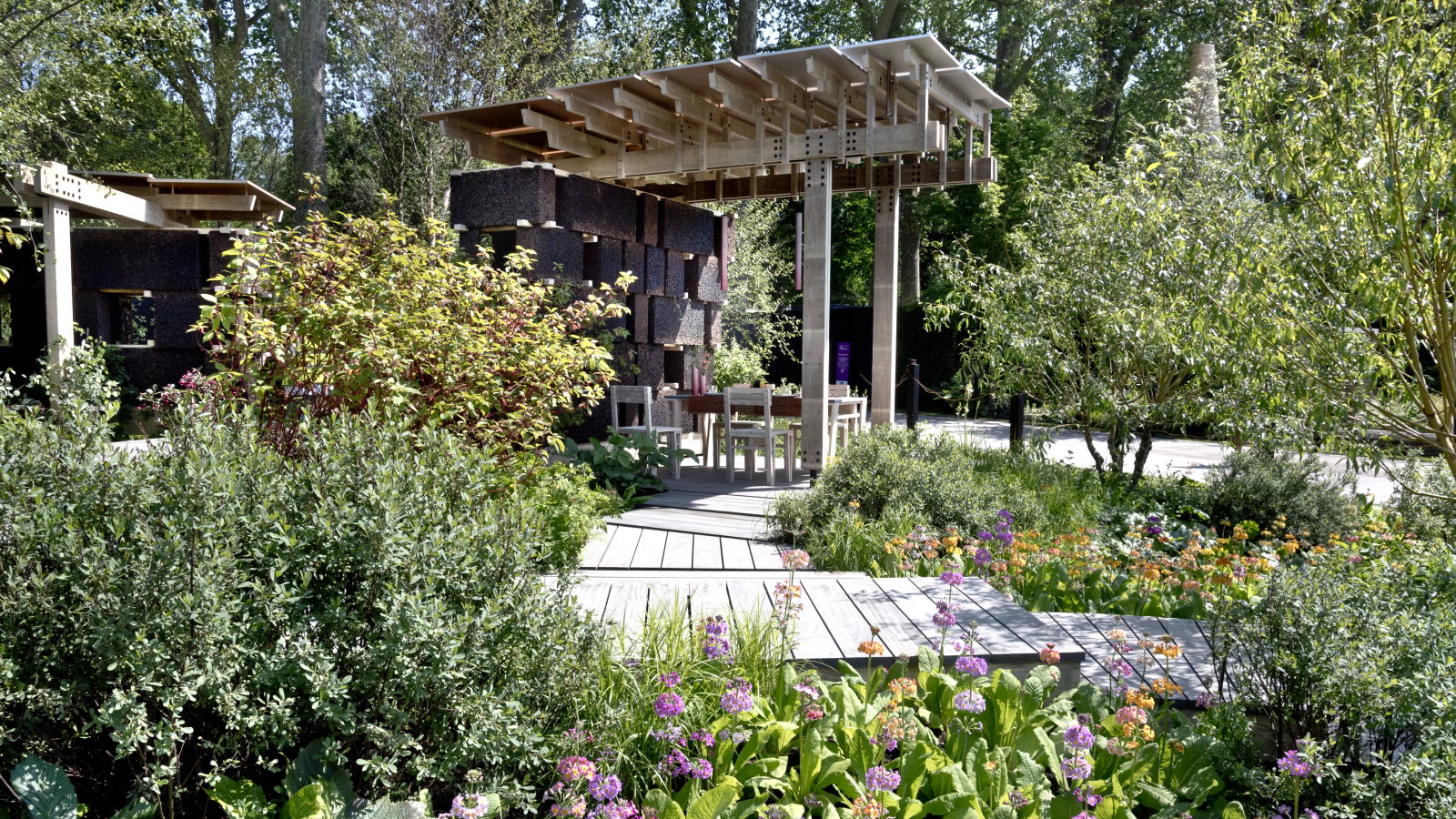 A show garden with shrubs and primulas alongside a wooden walkway and a large pergola with a dining set
