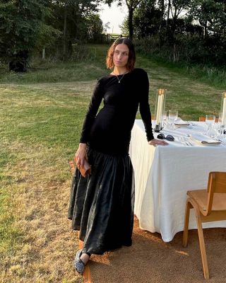 Influencer Erika Boldrin poses next to an outdoor dining table in Somerset, England wearing a black long-sleeved drop-waist dress and studded Mary Jane ballet flats