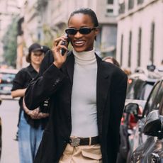 Woman on the phone wearing a white turtleneck, black blazer, and sunglasses 