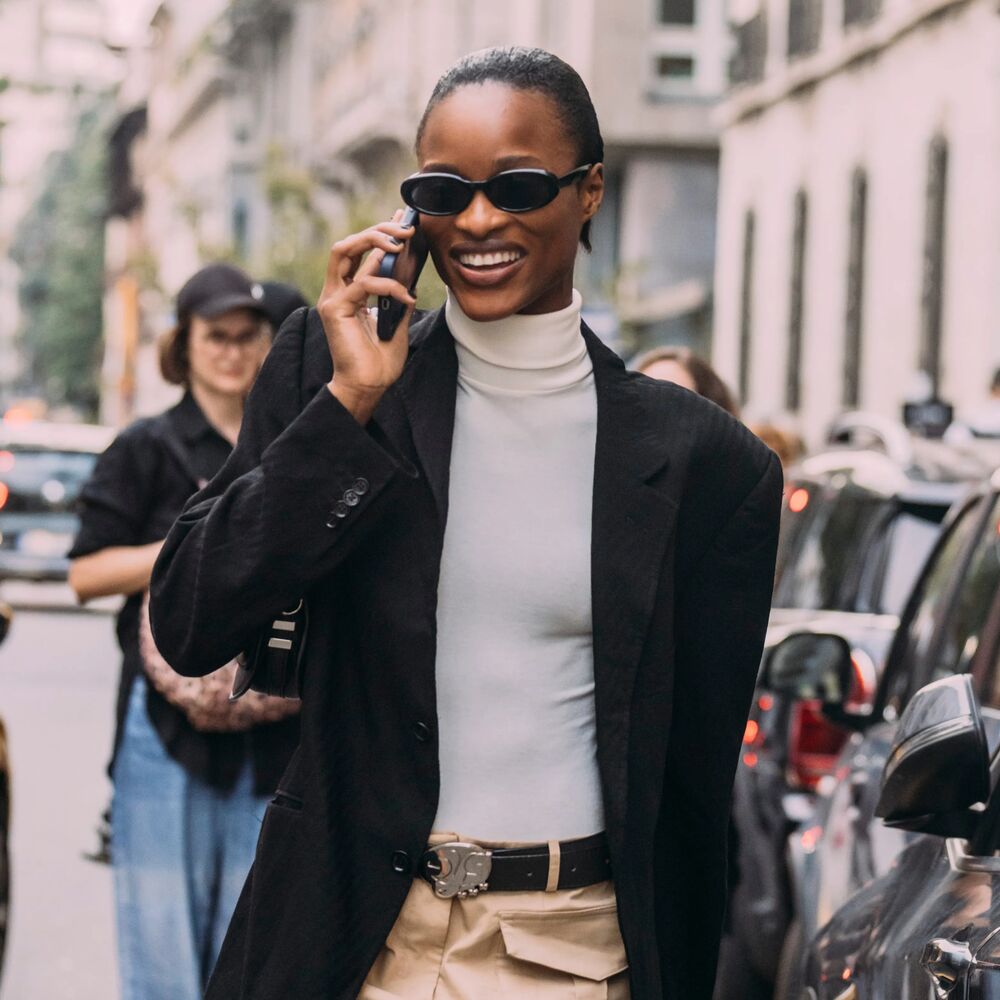 Woman on the phone wearing a white turtleneck, black blazer, and sunglasses 