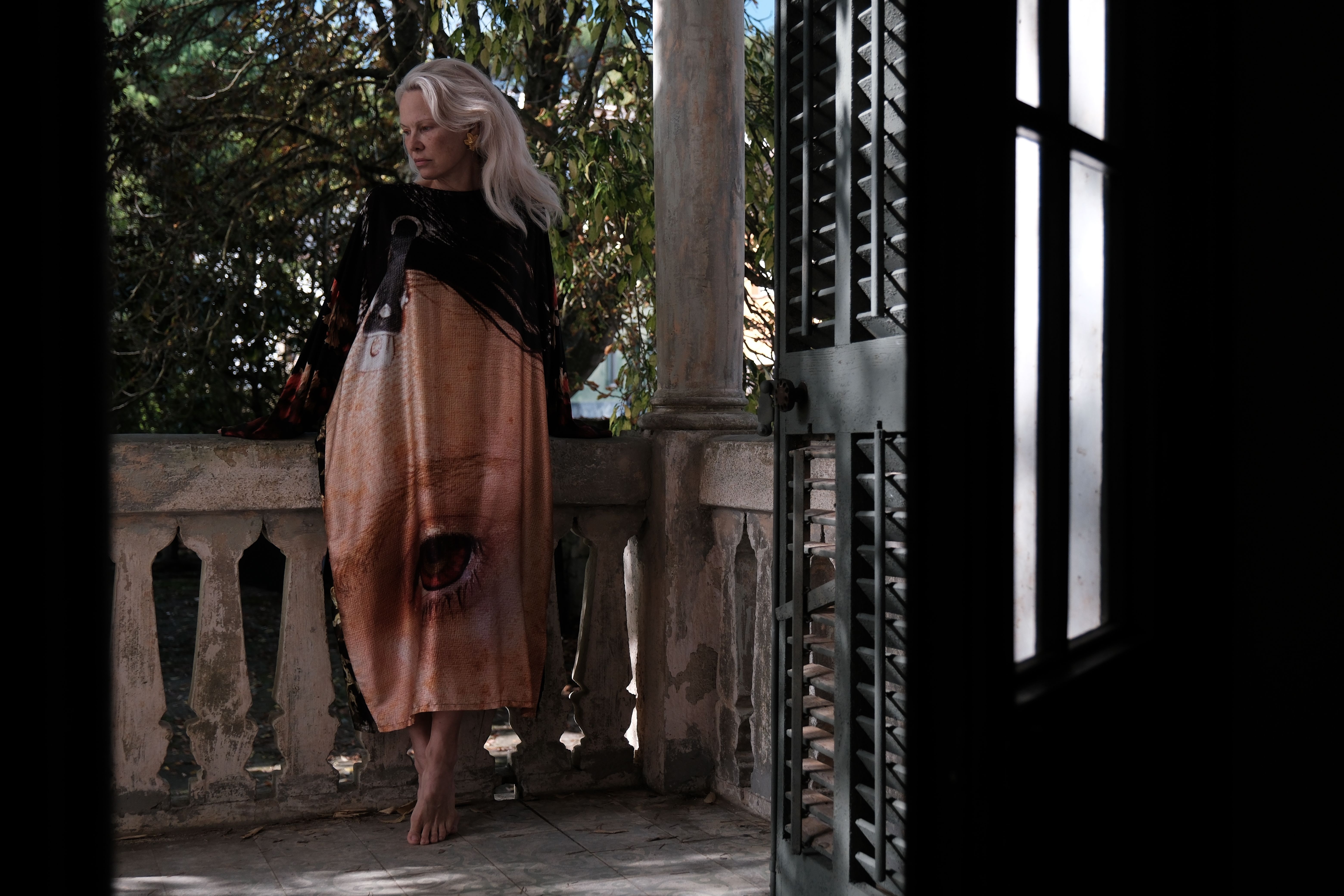 A blonde woman with voluminous blonde hair stands on an antique balcony dressed in a long-sleeved maxi dress with an eye drawn on it while looking pensive.