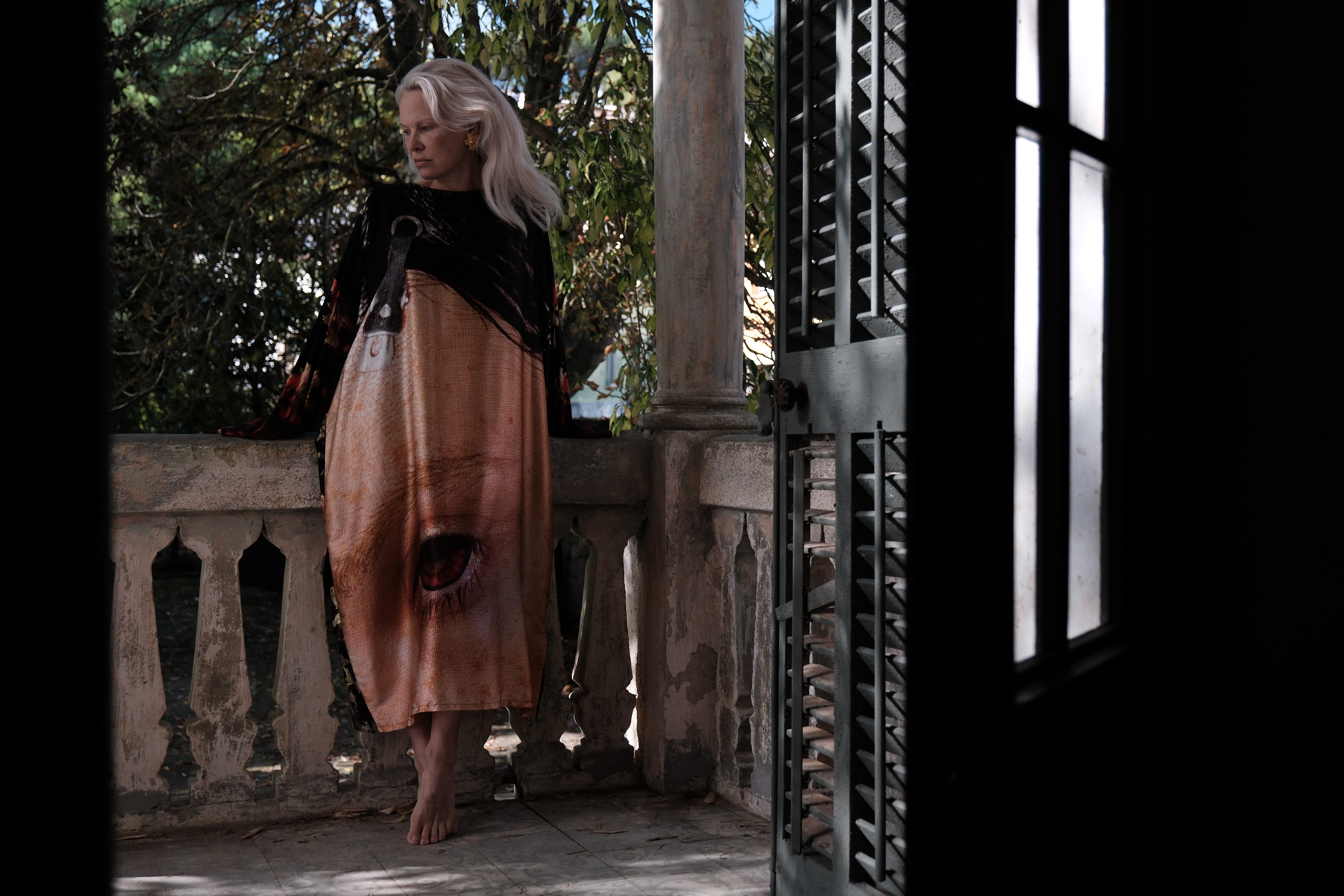 A blonde woman with voluminous blonde hair stands on an antique balcony dressed in a long-sleeved maxi dress with an eye drawn on it while looking pensive.
