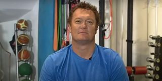 Luc Longley being interviewed in his workout room with basketballs and weights in the background.
