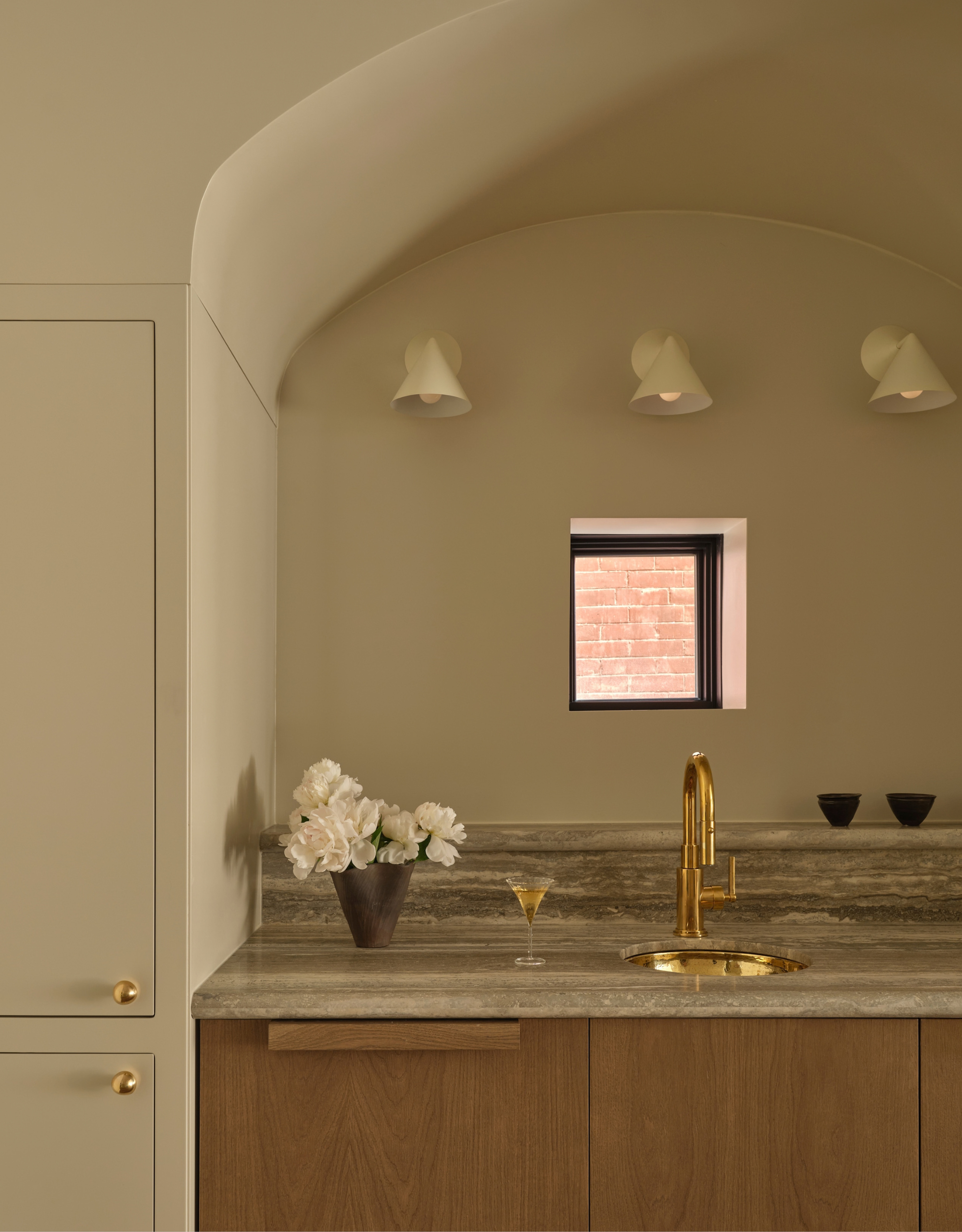 A kitchen with three conical wall sconces, a vase of flowers, a martini glass, a gold sink, and a pair of cups