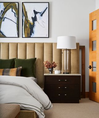 bedroom with white walls, abstract artwork in two frames on walls, long fabric headboard with black nightstand and plush neutral bedding with dark green cushions