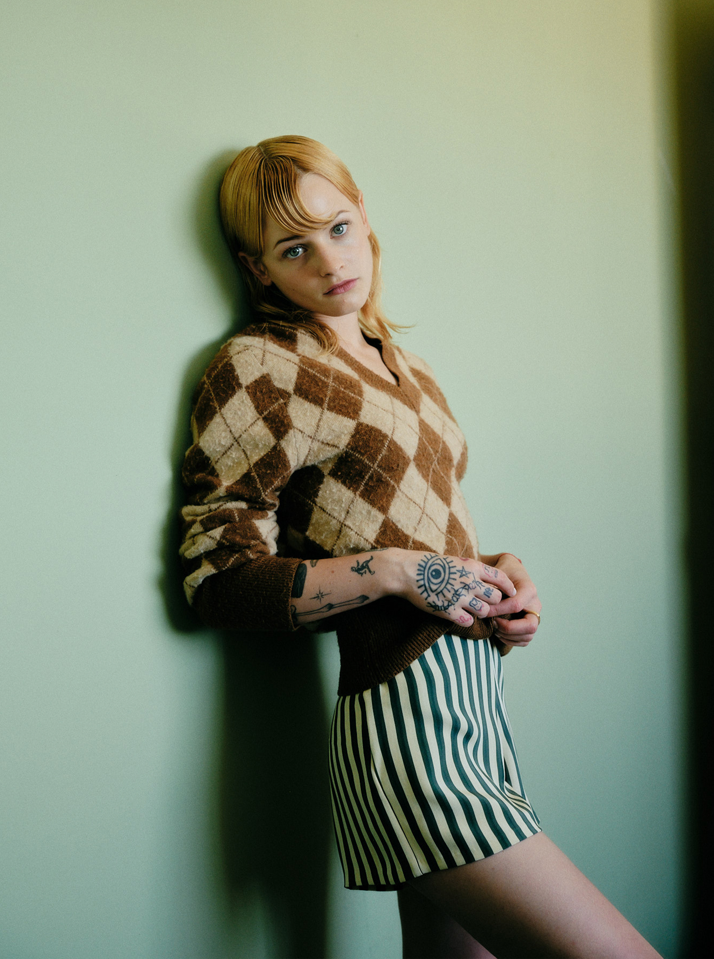 Esther McGregor leans up against a light green wall. She is wearing a brown and tan Coach argyle sweater and Bode blue and white stripe silk shorts.