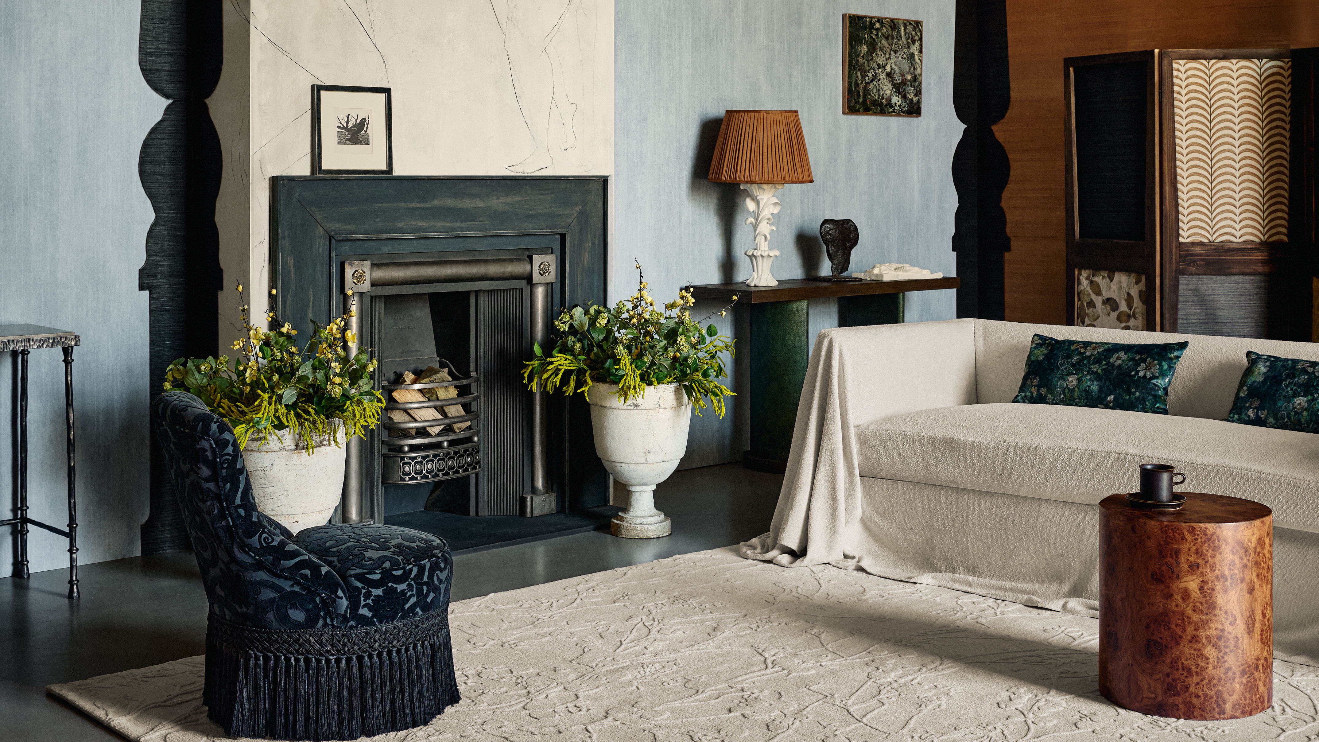 An elegant, textured living room features a dark fireplace flanked by two large stone urns filled with yellow flowers. To the left, a navy blue velvet chair with a deep fringe sits on a large, neutral-toned patterned rug. To the right, a white textured sofa with dark green floral pillows is positioned behind a round wood burl side table, while a console table in the background displays a white sculptural lamp with a pleated brown shade against light blue walls.