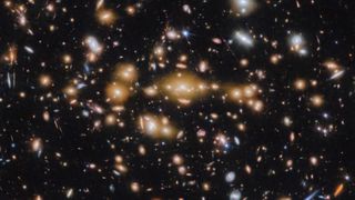 A series of glistening, golden dots in the center of the image show an ancient galaxy glowing in the early universe, surrounding by countless bright stars and galaxies