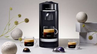 En Nespresso Vertuo Next er omgivet af kaffekapsler på et lyst bord.