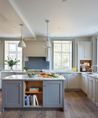 A kitchen with a gray kitchen island with colorful vegetables on top and books on the shelf, two cream pendant lights above it, two large windows on the wall, and cream cabinets to the back
