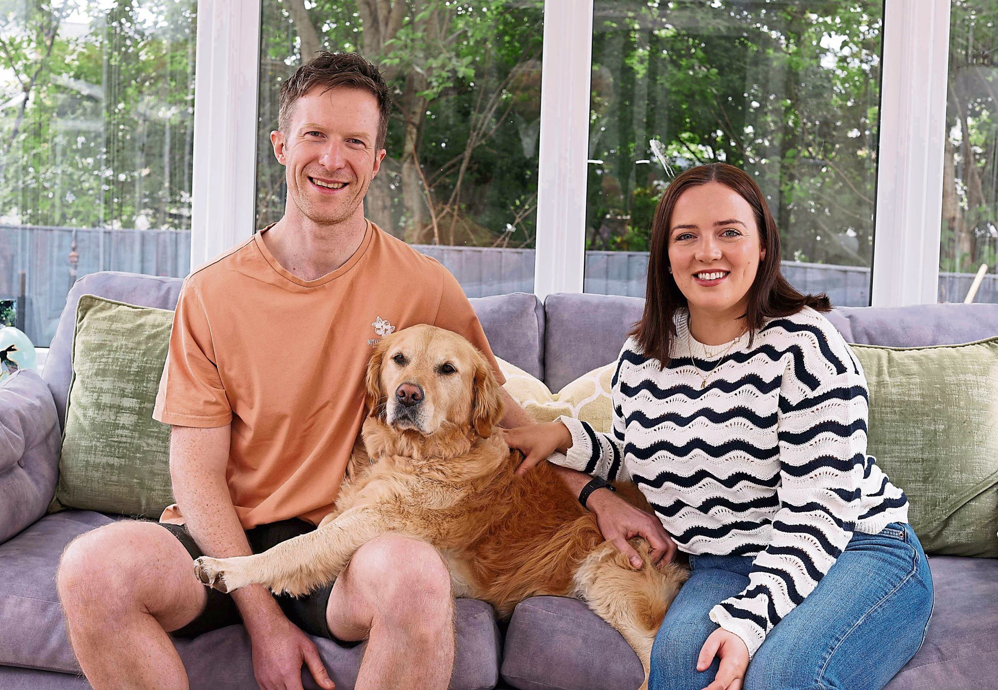 A Golden Retriever sits on a sofa between two adults in a bright conservatory, resting her paw on one person&amp;rsquo;s knee while both gently hold and stroke her. The dog looks calm and content as the pair smile towards the camera.