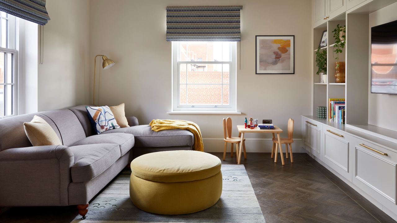 Living room with grey sofa, yellow ottoman, and large windows with blinds