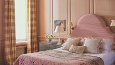 Bedroom with warm pink walls, upholstered pink bed with curved headboard, a pink quilt, striped throw pillows and white bed linen, a nightstand with a horse painting above it and plaid yellow curtains