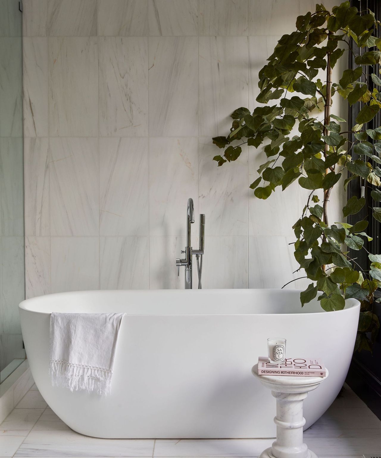 A white freestanding bath in a light tiled bathroom paired with an indoor tree and a rustic white stool