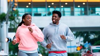 Man and woman talk while jogging in a city. They are both wearing tracksuits.