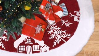 A red tree skirt with trees, reindeer and a house embroidered on it.