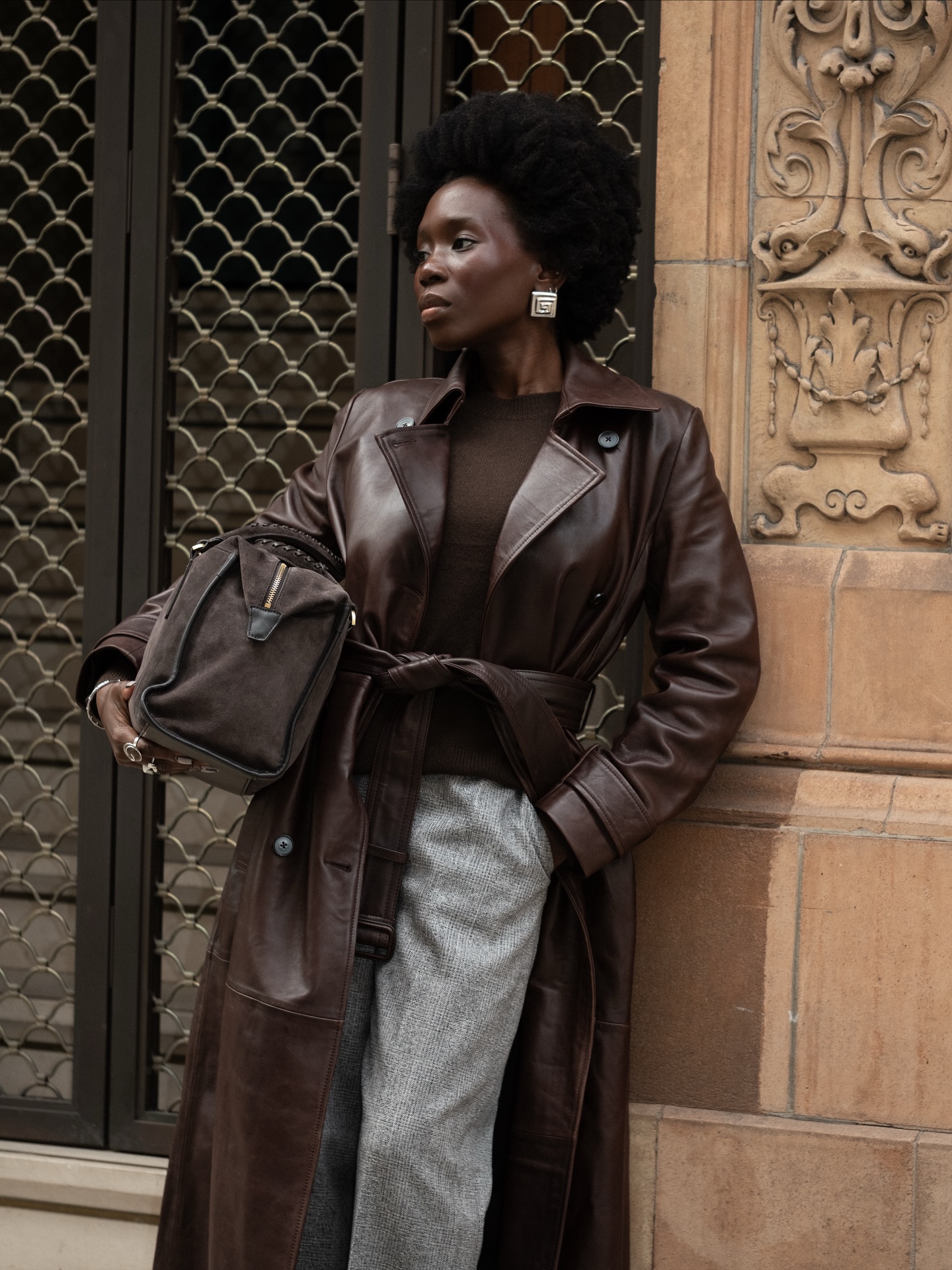 An image of an influencer wearing a brown leather coat, a brown shirt and grey trousers carrying a brown suede bag